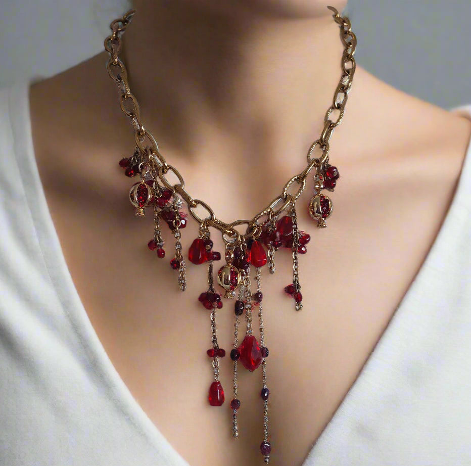 A person wearing a white v-neck top showcasing a gold-plated stainless steel bib necklace with pomegranate-shaped natural garnet and ruby beads along with glass beads.